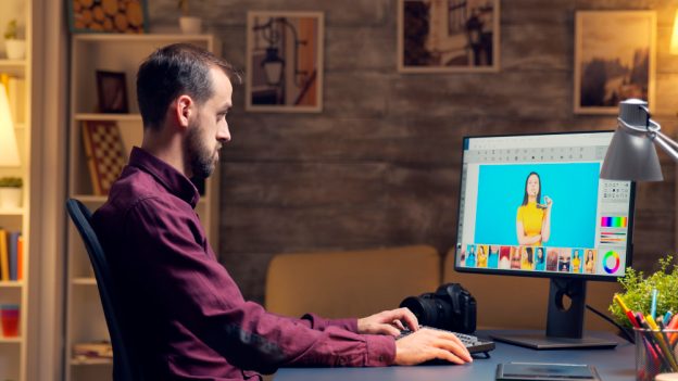 Video editor working on dual monitors with editing software, while a woman sits in the background on a couch, talking on the phone.